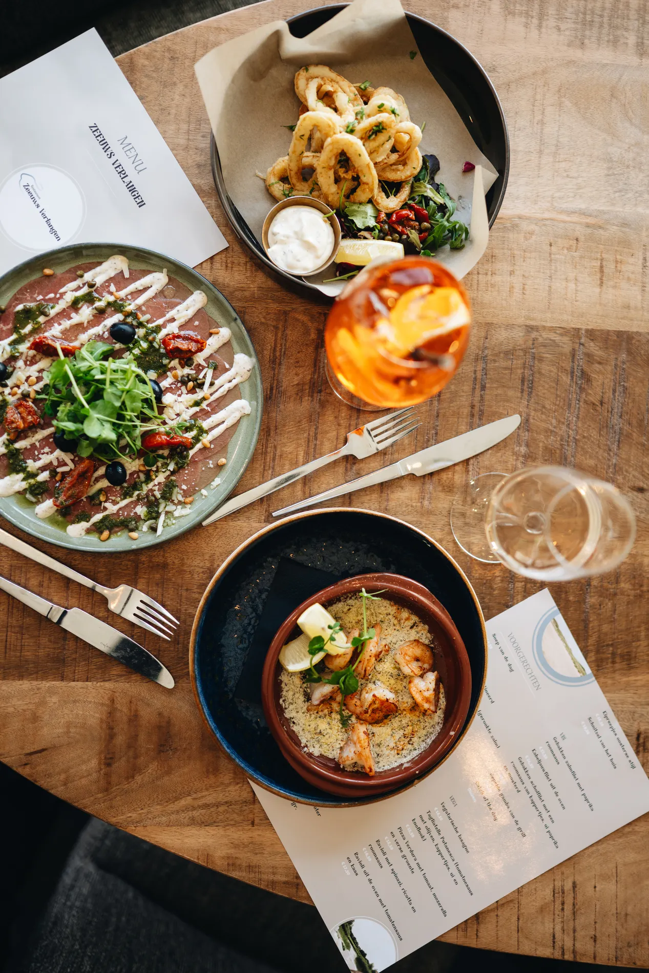 Bovenaanzicht van een houten tafel vol met gerechten, waaronder een bord carpaccio met rucola, een schaal gefrituurde calamares, knoflookgarnalen, een Aperol Spritz cocktail en de menukaart van het restaurant.